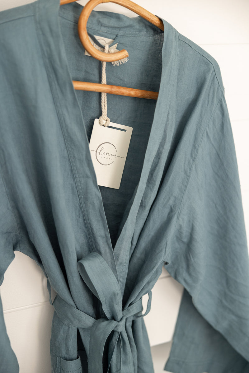 Linen Label Dust Denim Blue French Linen Bathrobe Kimono Robe hanging on a detailed cane hanger - looking luxurious and aesthetically pleasing in a sage green, perfect for ultimate comfort and relaxation. Crafted with 100% French Linen that grows softer with each wash