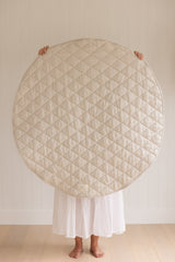 A women holding up a Linen Label Pure French Linen quilted padded play mat folded showing the Natural Oat colour side, she hold it over her face to show the size. 