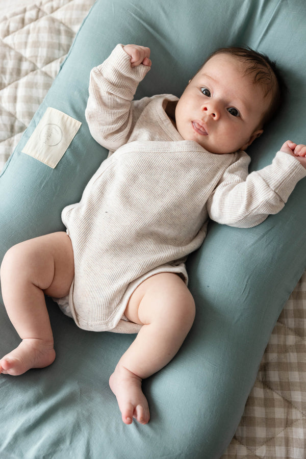 A baby looking content laying on Linen Label baby nest lounger with Dugg Egg Blue French Linen Cover, providing a lightweight, breathable, and hypoallergenic sanctuary for your little one.