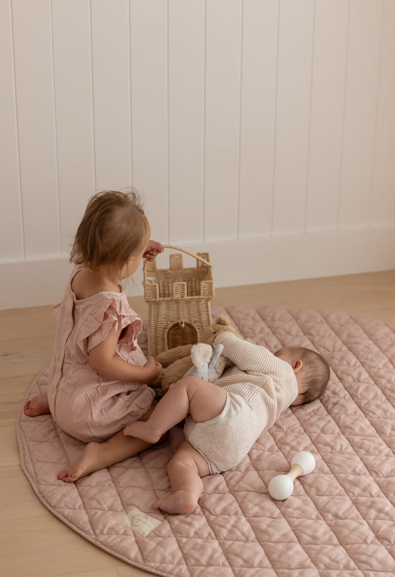 two babies playing, one sitting and one laying both on Linen Label Pure French Linen quilted padded play mat in Dusty Rose Pink colour and the other Gingham check