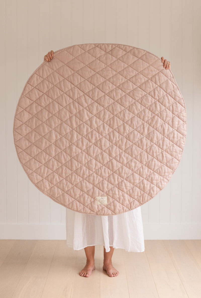 A women holding up a Linen Label Pure French Linen quilted padded play mat folded showing the Dusty Rose Pink colour side, she hold it over her face to show the size.