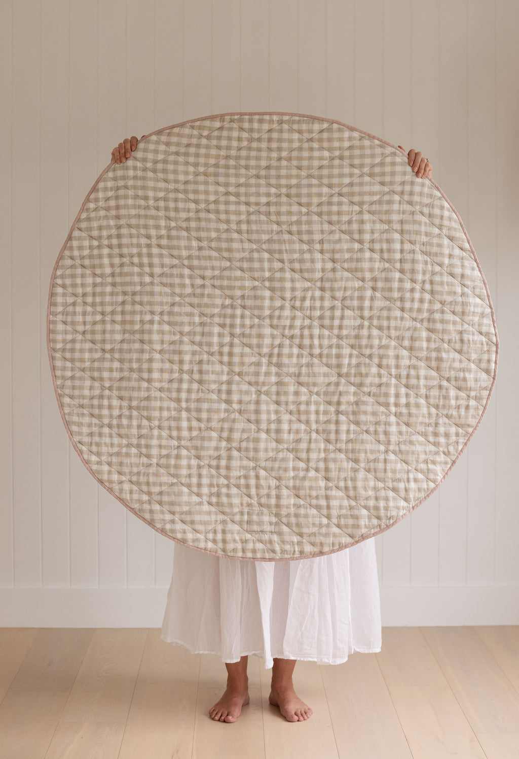 A women holding up a Linen Label Pure French Linen quilted padded play mat folded showing the reverse side of Natural Oat gingham colour side with Dusty Rose Pink gingham binding, she hold it over her face and baby to show the size.