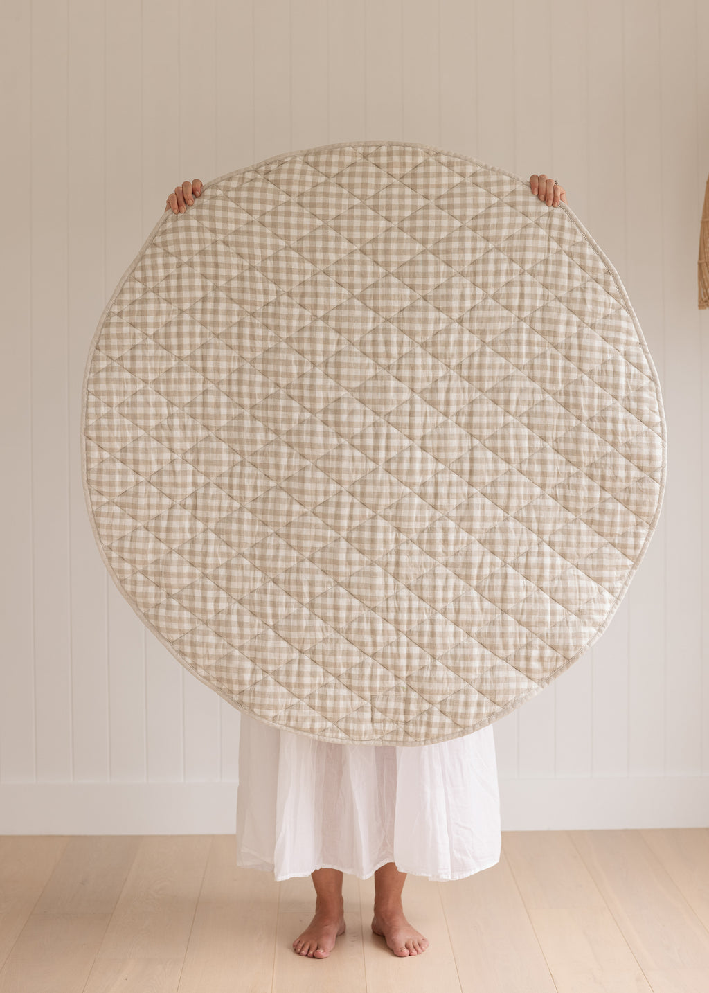 A women holding up a Linen Label Pure French Linen quilted padded play mat folded showing the reverse side of Natural Oat gingham colour side, she hold it over her face and baby to show the size. 
