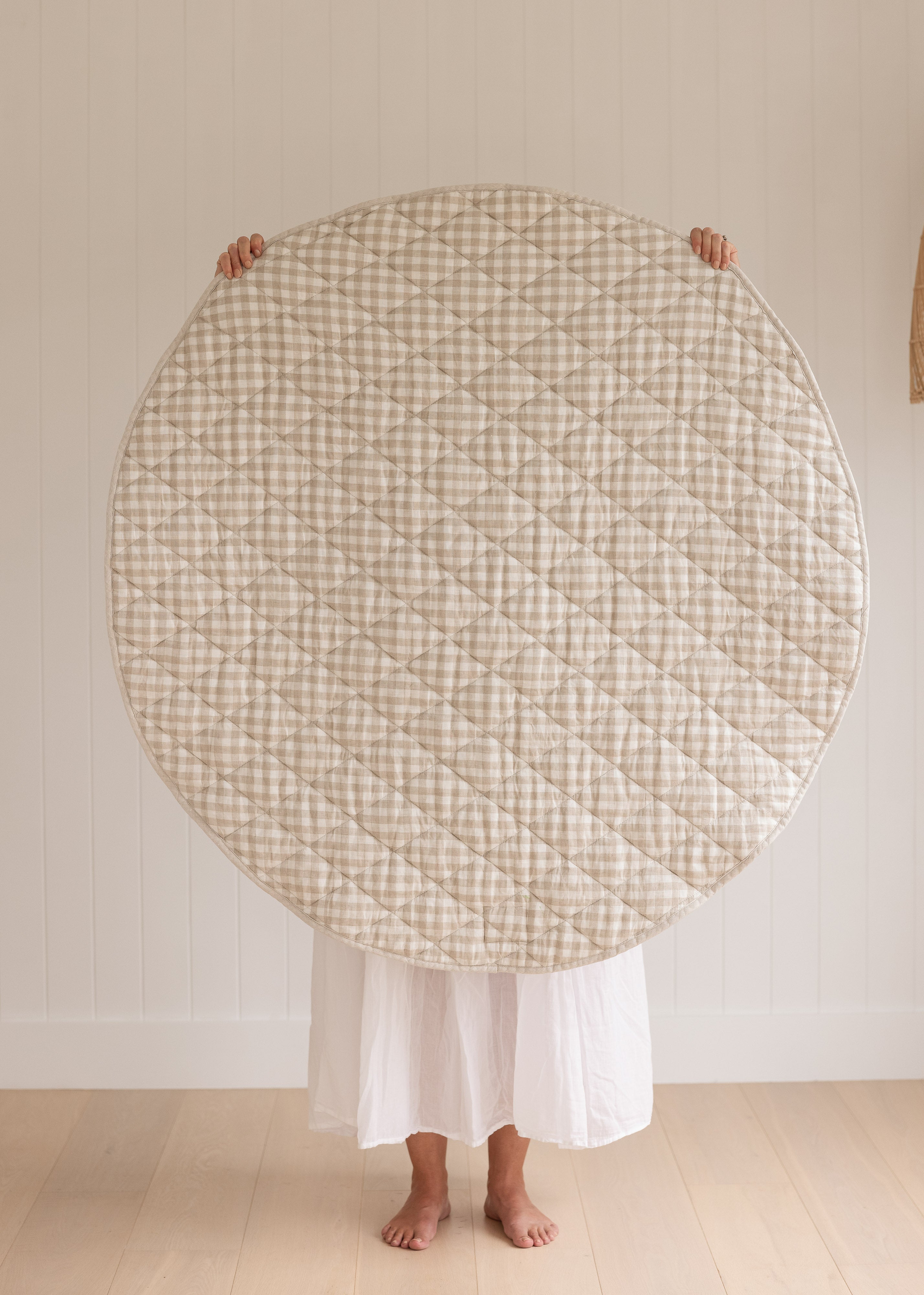 A women holding up a Linen Label Pure French Linen quilted padded play mat folded showing the reverse side of Natural Oat gingham colour side, she hold it over her face and baby to show the size. 