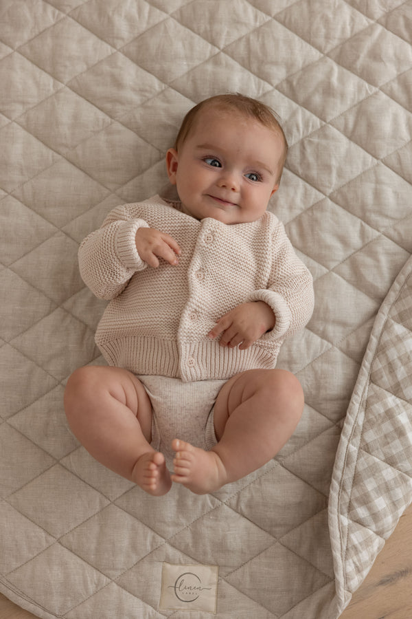 A close photo of a baby kicking around and playing on Linen Label Pure French Linen showing the quilted padded detail of the play mat. The edge is folded showing both sides, one side Natural Oat beige and the other side is Natural Gingham check