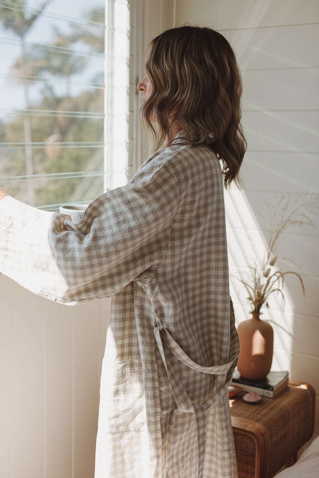 Woman looking out at the morning sun in her Linen Label Natural Oat gingham French Linen Bathrobe Kimono - looking luxurious in her soothing sage green linen robe, perfect for ultimate comfort and relaxation. Crafted with 100% French Linen that grows softer with each wash. Ideal as a dressing gown, bathrobe, or pyjamas. 