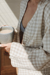 Mother dressed in Linen Label Natural Oat Gingham  French Linen Bathrobe Kimono Robe enjoys some downtime with a coffee in hand. looking stylish and comfortable around the home. Showing Linen Label Robes are perfect for ultimate comfort and relaxation. Crafted with 100% French Linen that grows softer with each wash