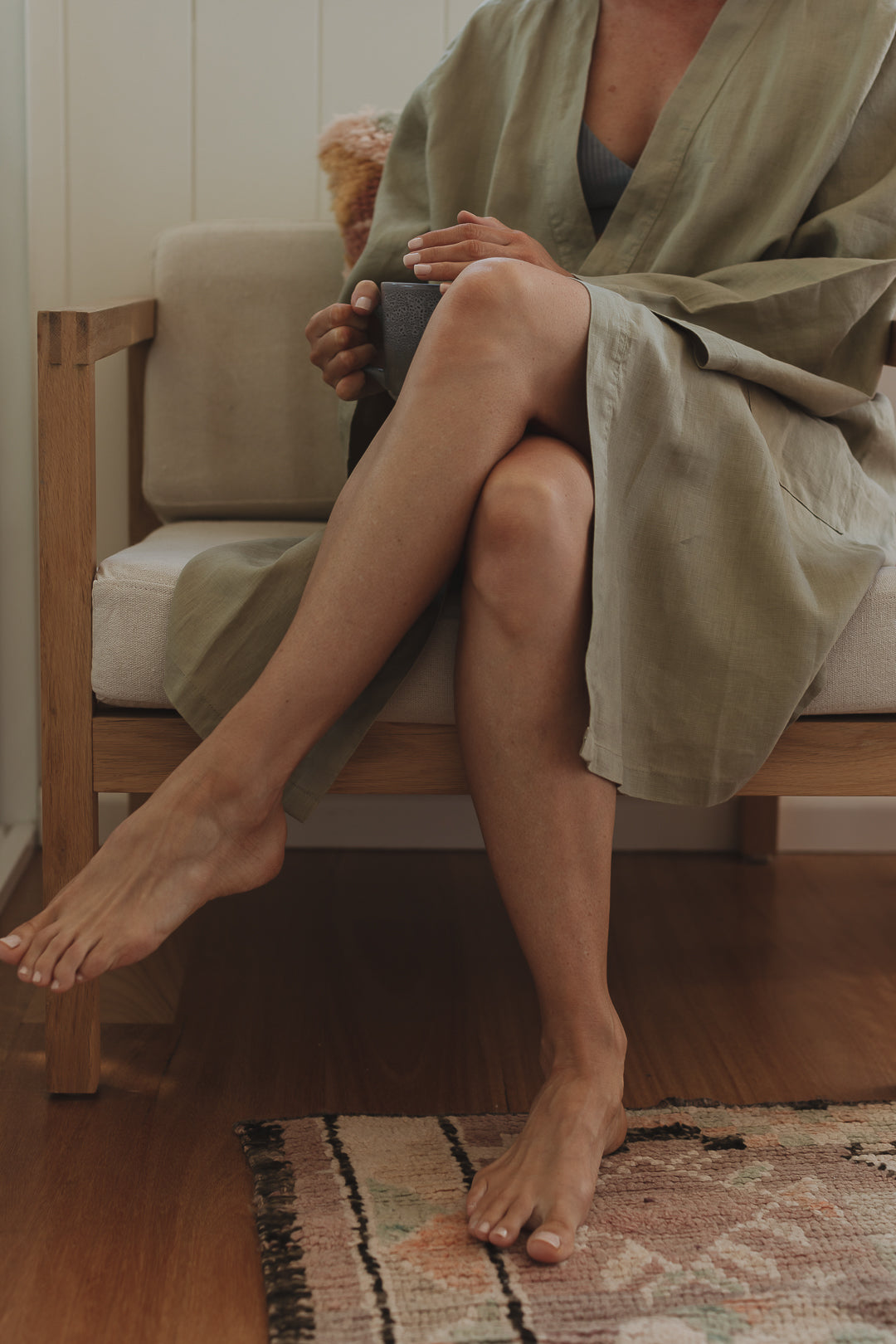 Mother dressed in Linen Label Soft Sage Green French Linen Bathrobe Kimono Robe her baby sleeping while she enjoys some downtime with a coffee in hand. looking stylish and comfortable around the home. Showing Linen Label Robes are perfect for ultimate comfort and relaxation. Crafted with 100% French Linen that grows softer with each wash