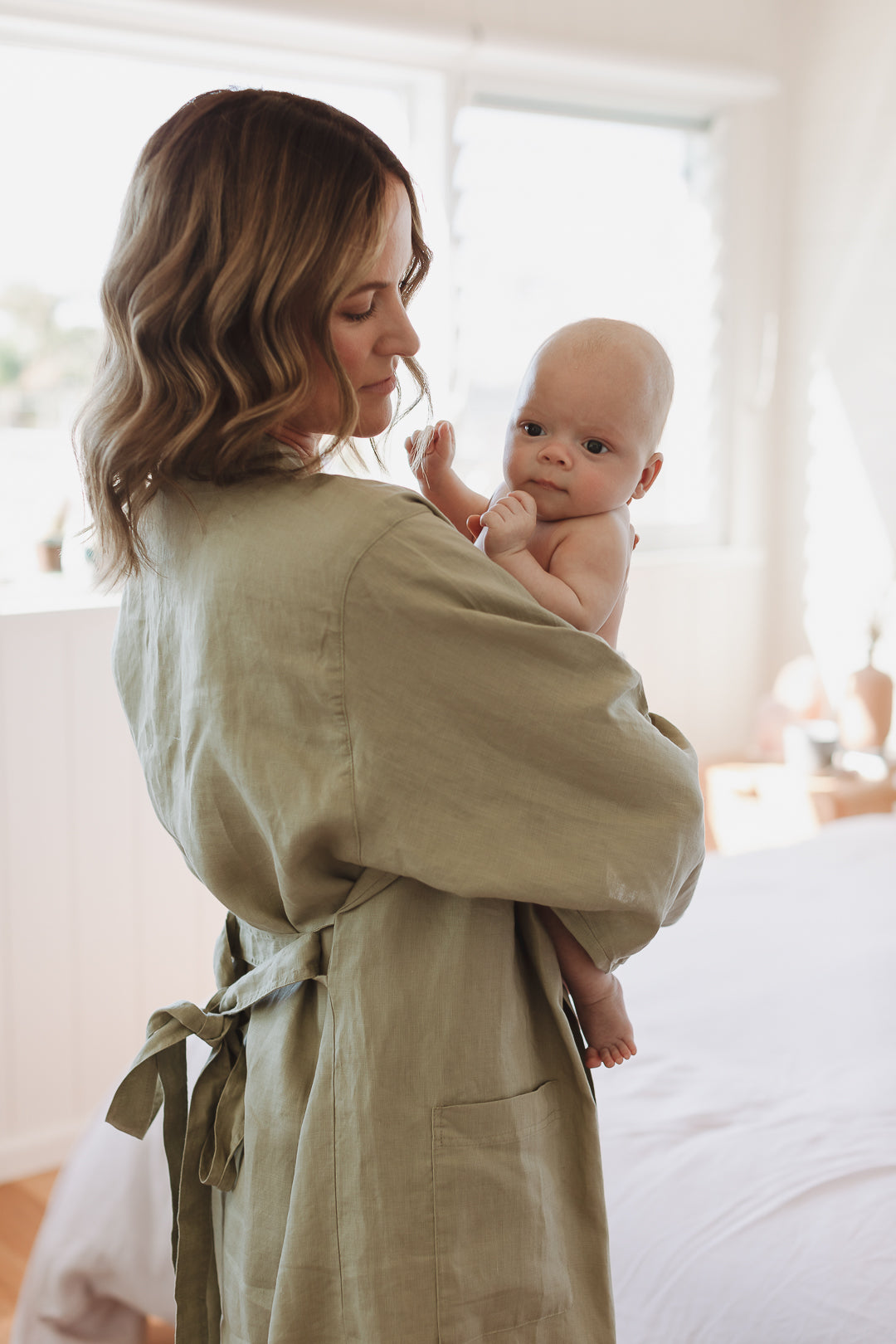 Mother dressed in Linen Label Soft Sage Green French Linen Bathrobe Kimono Robe holding a newborn baby over her shoulder after feeding. looking stylish and comfortable around the home. Showing Linen Label Robes are perfect for ultimate comfort and relaxation. Crafted with 100% French Linen that grows softer with each wash. 