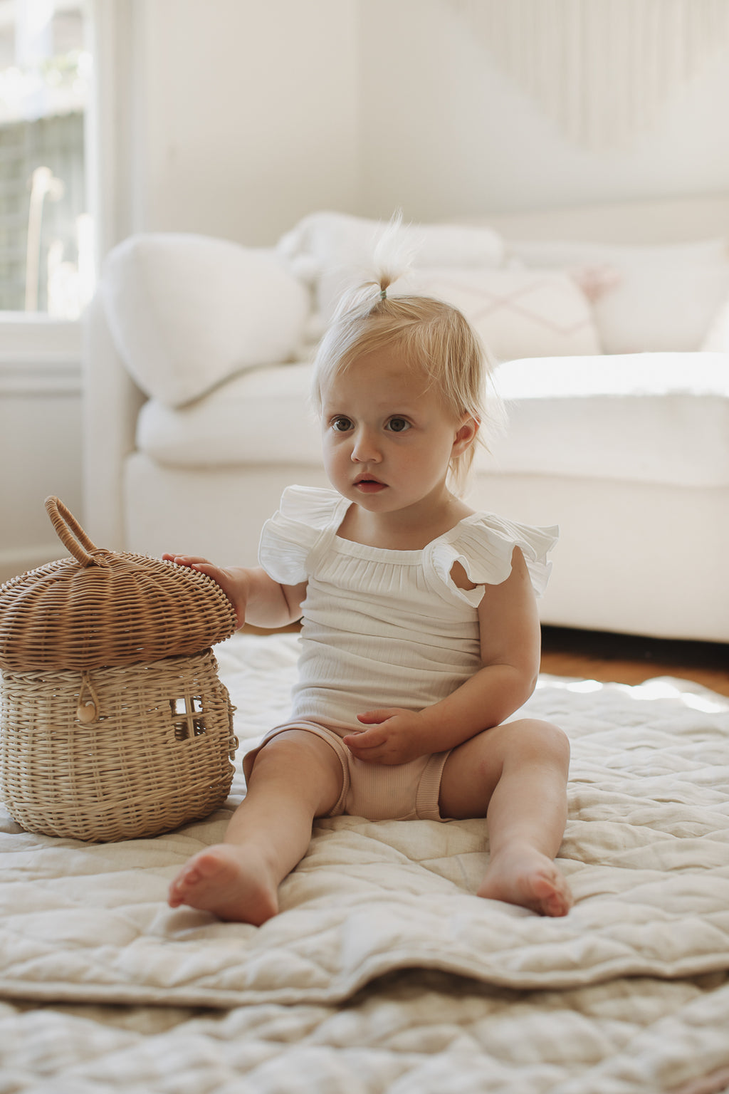 Baby girl sitting up playing with basket on aLinen Label Pure French Linen quilted padded play mat folded showing both sides, one Natural Oat colour and the other Gingham check