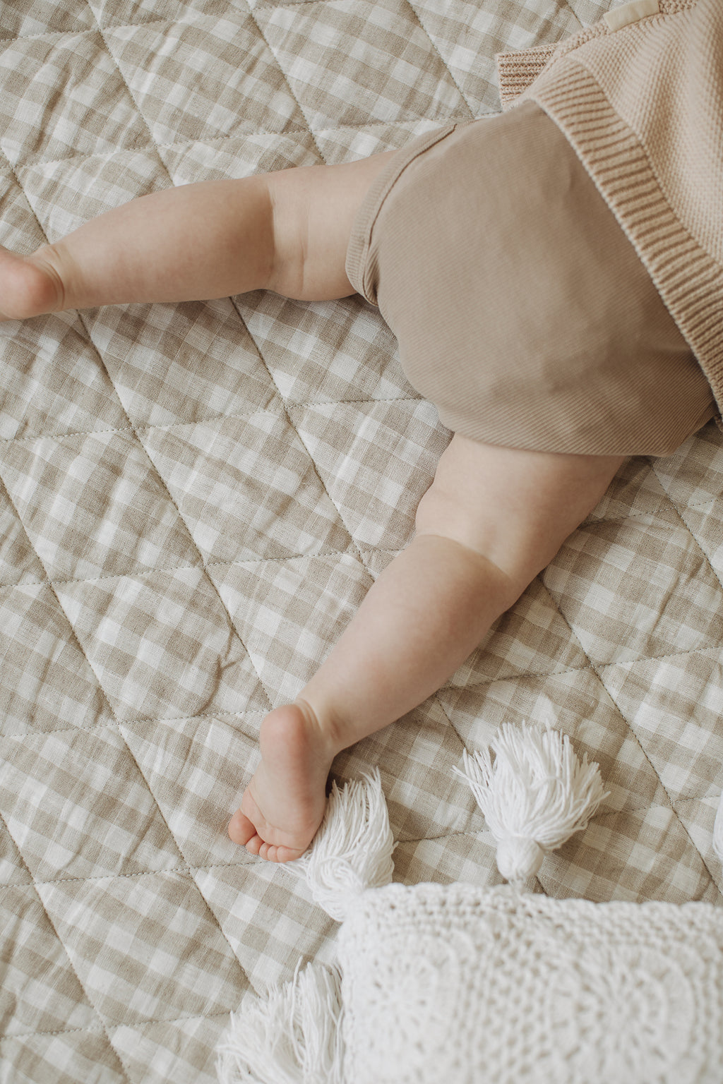 ALittle baby feet and leg tummy time on the Linen Label pure French Linen quilted padded play mat folded showing the softness of the Natural Oat colour and the other Gingham check
