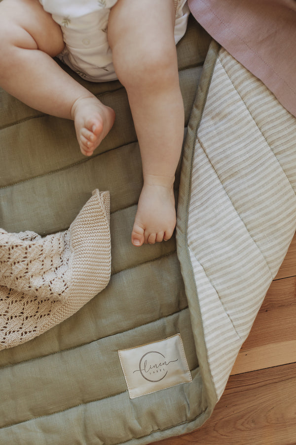 A close photo of a baby feet and legs playing on Linen Label Pure French Linen showing the quilted padded detail of the play mat. The edge is folded showing both sides, one soft Sage Green colour and the other pinstripe natural and white colour